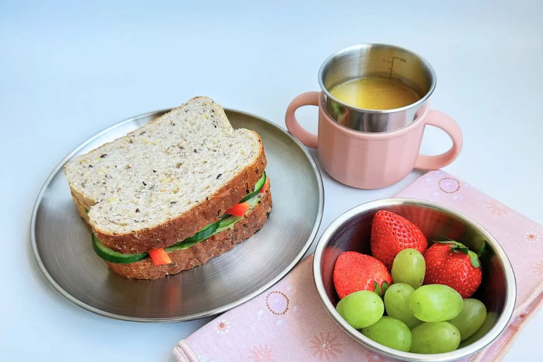 45 degree angle view of kids stainless steel plate with food, bowl with fresh fruits, and sippy cup with a drink, showcasing eco-friendly Greenvyne tableware.