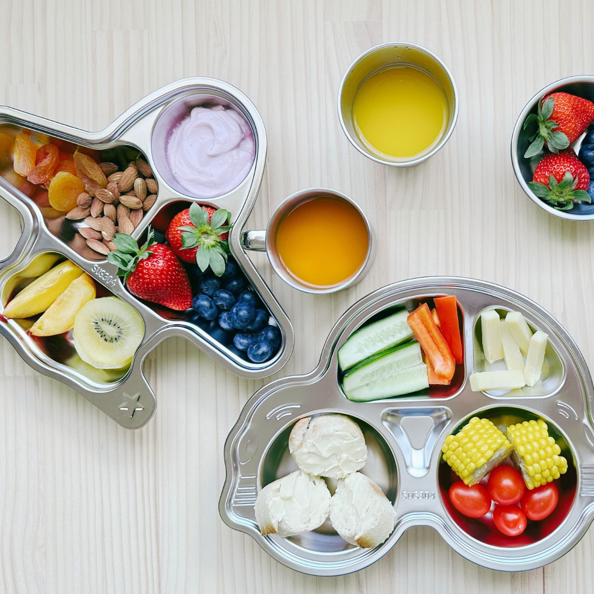 Top view of a stainless steel car plate and plane plate filled with food, a 250ml mug with juice, a 300ml cup with juice, and a 10cm bowl with fruits on a table, showcasing a complete eco-friendly mealtime setup.