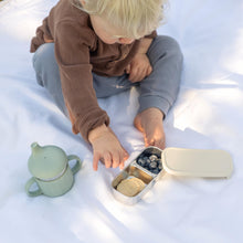 Toddler using Greenvyne snack box stainless steel with leak resistant Oatmeal silicone sleeve, filled with fruits and snacks.