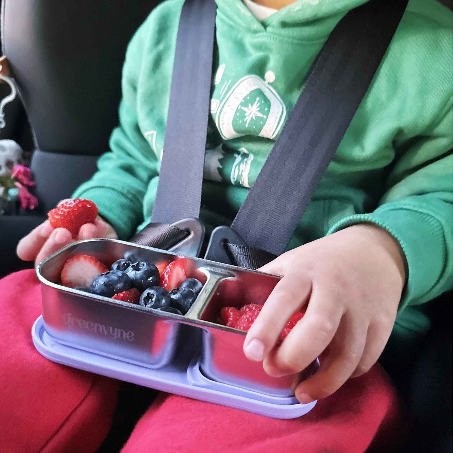 A child in a car seat holding a bento snack box with fruits, showcasing versatility - Greenvyne.