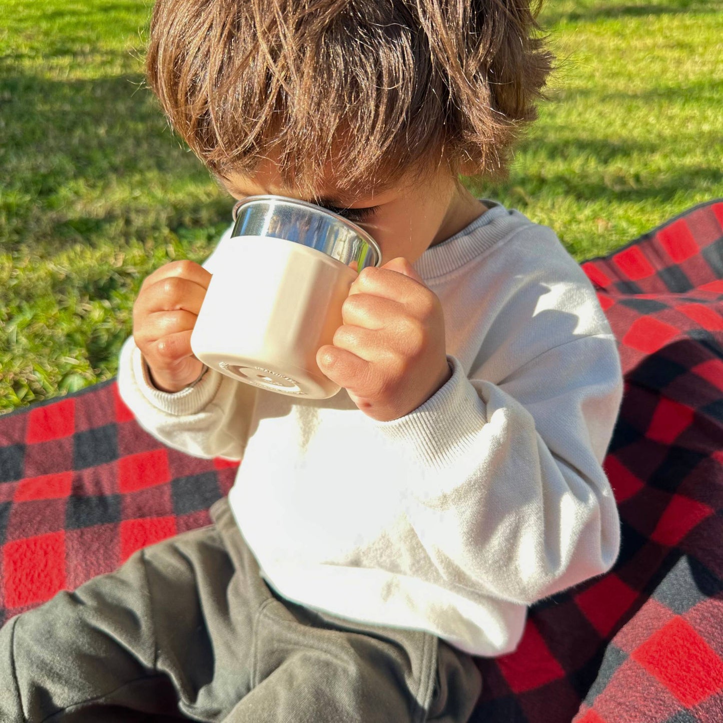 Child drinking from a Greenvyne stainless steel sippy cup outdoors on a picnic blanket.