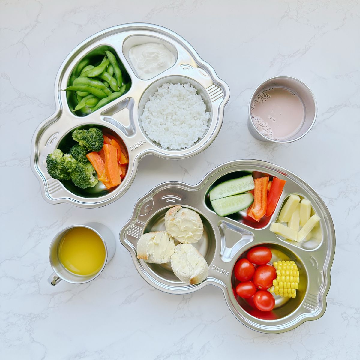 Top view of two stainless steel car plates filled with food, a 250ml mug with juice, and a 300ml cup with juice on a table, showcasing an eco-friendly and kid-friendly mealtime setup.