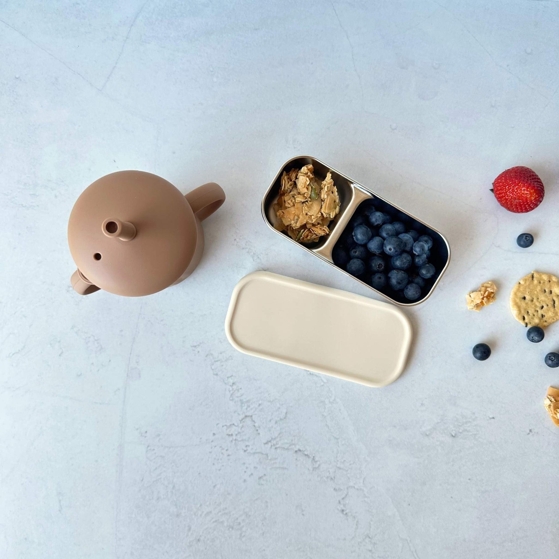 Greenvyne toddler two compartment snack boxes stainless steel with oatmeal colour silicone lid, filled with snacks and a brown colour stainless steel baby & toddler sippy cup on table.