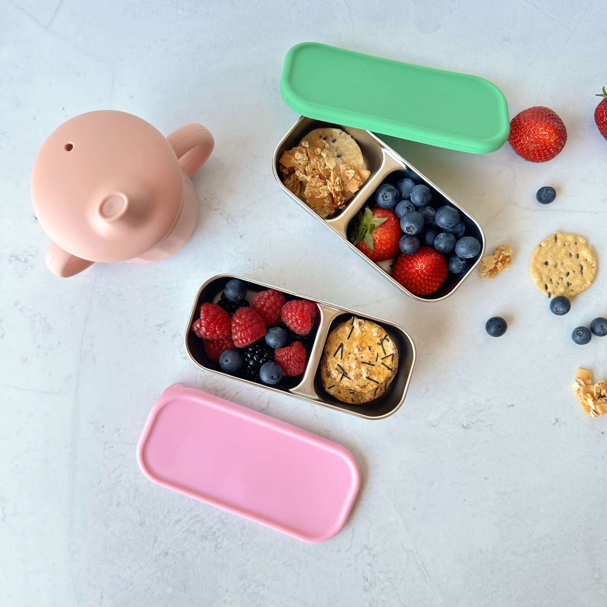 Greenvyne toddler two compartment snack boxes stainless steel with pink and green colour silicone lids, filled with fruits and snacks and a pink toddler sippy cup.