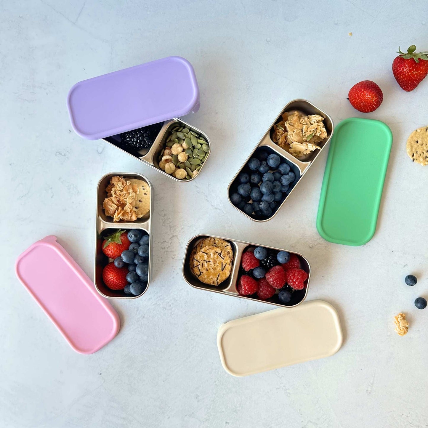 Greenvyne Kids two compartment snack box stainless steel with purple, green, pink and cream colour silicone lids, filled with fruits and snacks.