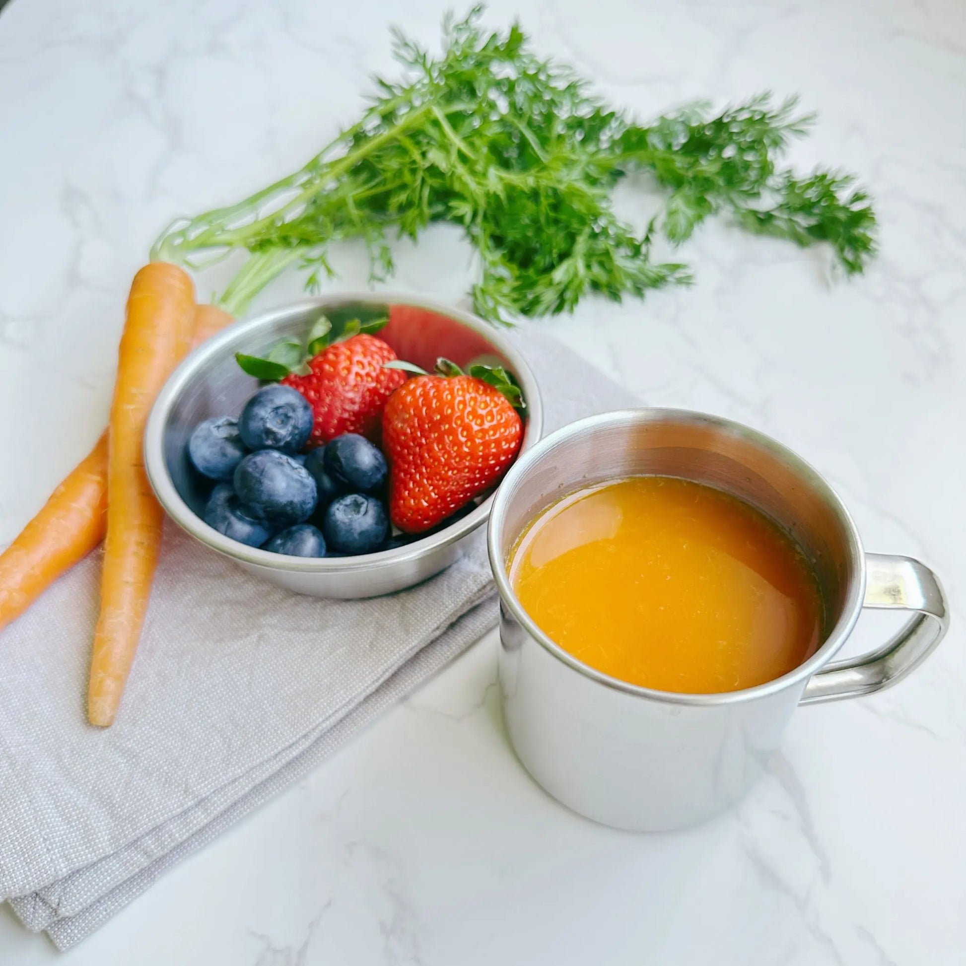 45-degree angle view of a 250ml stainless steel mug filled with juice, a 10cm stainless steel bowl with fruits, and fresh carrots on a table, showcasing an eco-friendly and versatile dining setup.