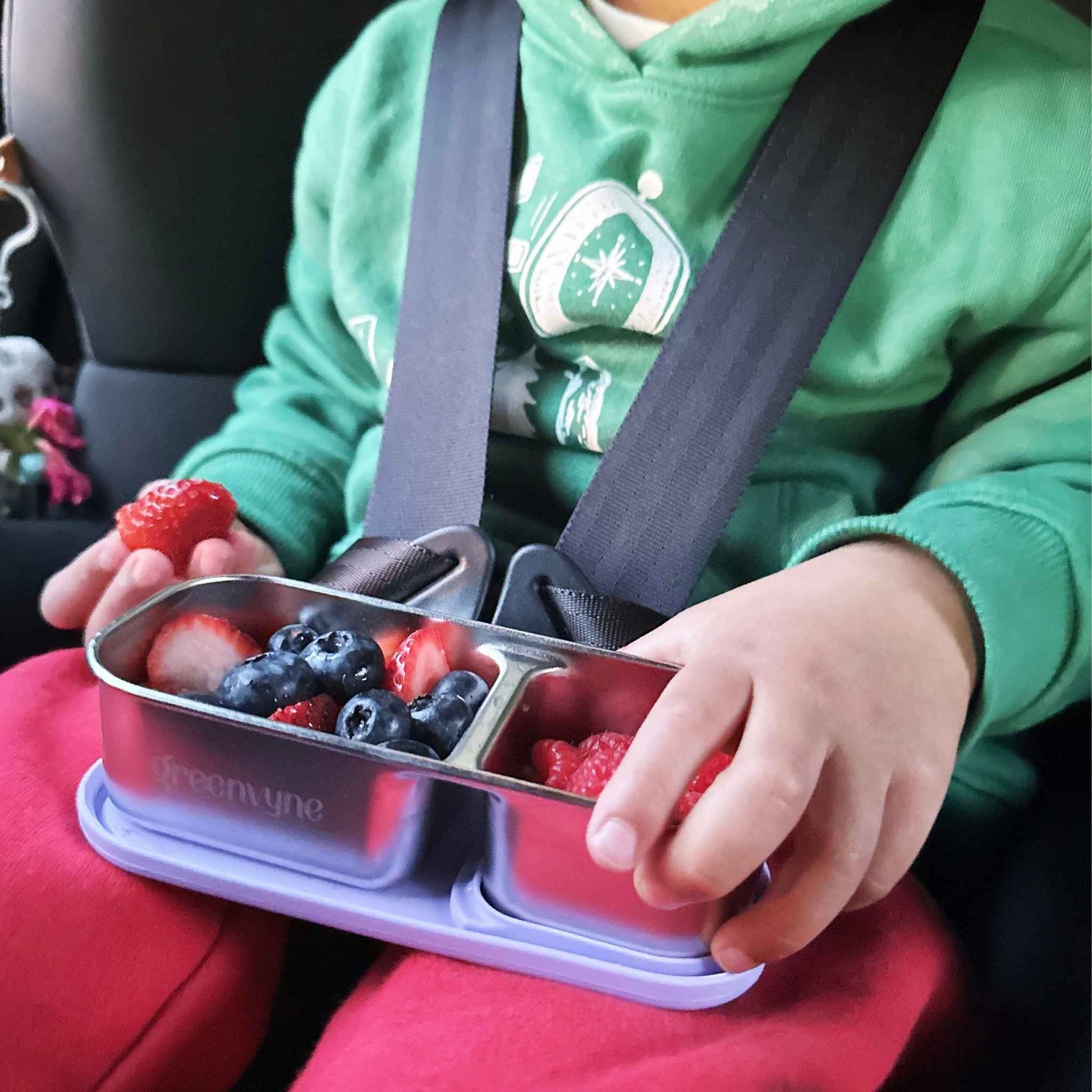 A child in a car seat holding a bento snack box with fruits, showcasing versatility - Greenvyne.