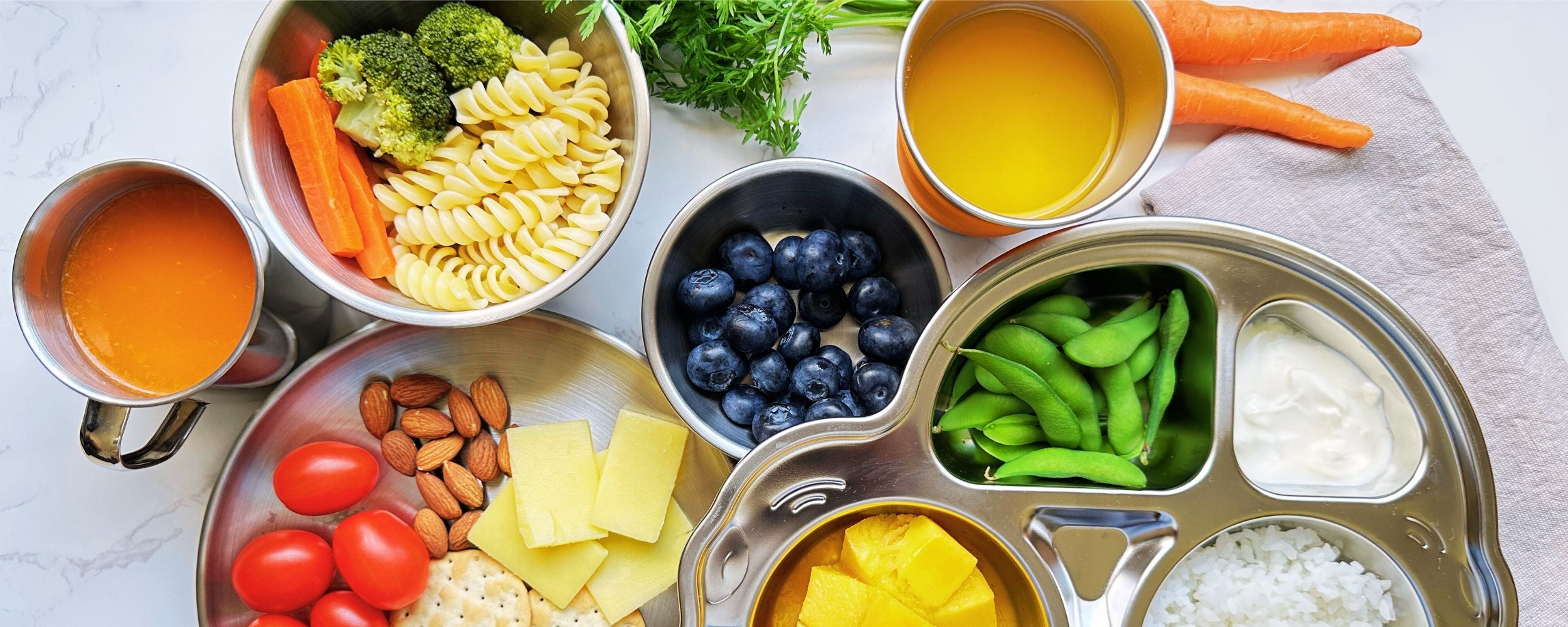 Cropped top view of a stainless steel mug and cup with juice, two bowls, a car-shaped plate with food and fruits, a meal plate with snacks, and fresh carrots placed on a table, showcasing a complete eco-friendly dining setup.